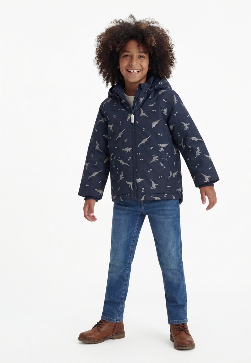 Kind met krullend haar, gekleed in een marineblauw jack met dinosaurussen, blauwe spijkerbroek en bruine laarzen, staand en lachend tegen een witte achtergrond.