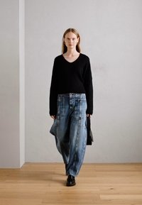 Suéter de punto negro con textura y escote en V, combinado con pantalones de mezclilla azul claro y sueltos que presentan grandes bolsillos. La modelo sostiene un bolso negro.