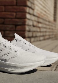 Coppia di scarpe da corsa atletiche bianche con trama a maglia e tre distintive strisce, posate su pavimentazione esterna vicino a un muro di mattoni.