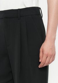 Die rechte Hand liegt neben einer schwarzen, hoch taillierten, gefalteten Hose mit Gürtelschlaufen, die mit einem weißen Oberteil getragen wird.