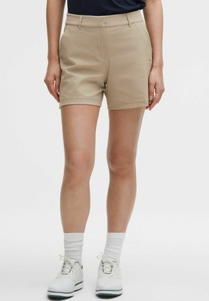 Mujer llevando pantalones cortos beige a medida, calcetines blancos y zapatillas blancas, de pie contra un fondo liso.