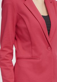 Blazer rose ajusté en tissu texturé, avec un col à revers cranté, une fermeture à un bouton et une poche latérale.