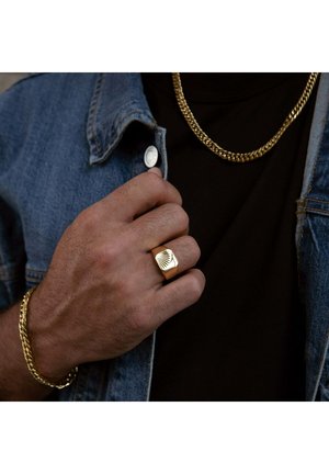 Bague en or avec un design géométrique texturé, portée à la main. Fini avec une surface lisse, associée à un bracelet et un collier en or épais.