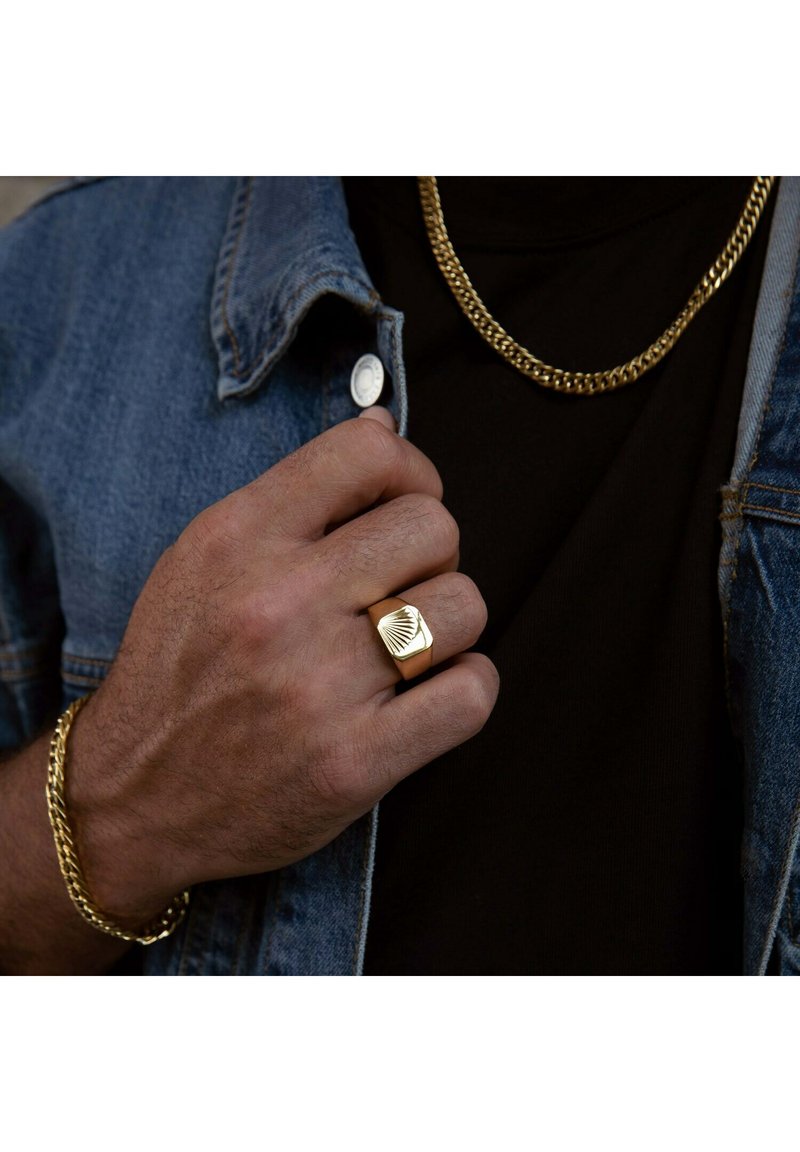 Gold ring with a textured geometric design, worn on a hand. Finished with a smooth surface, paired with a thick gold chain bracelet and necklace.