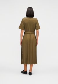 Ελιά πράσινο, κον τομάνικο φόρεμα με πλισέ φούστα και ζωνάκι στη μέση. Η πλάτη έχει ευθεία κοπή και πλαϊνές σχισμές για ευκολία στην κίνηση.