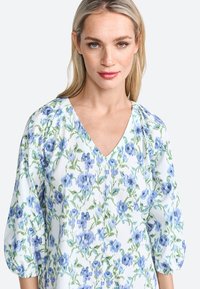 Blumenbluse mit V-Ausschnitt, puffigen Ärmeln und einem hellblauen Hintergrund mit grünen und blauen Blumenmustern. Weiche Stoffstruktur.