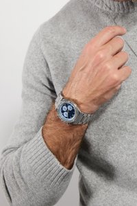 Reloj de acero inoxidable plateado con un dial redondo azul, tres subdiales, patrón texturizado y acentos de cristal en el bisel, llevado en una muñeca.
