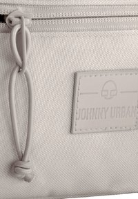 Detailní záběr na béžový látkový p Houstonu s zipem a smyčkovým tahem, který má přišitou obdélníkovou cedulku s logem a textem.