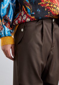 Bruine plooibroek met een gladde textuur, met een opening voor een zijzak en een metallic knoopaccent. Kleurrijke gebreide trui zichtbaar.