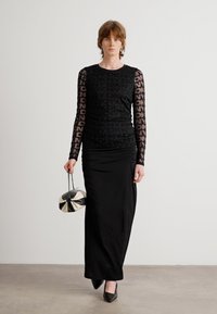 Schwarzes langärmliges Kleid mit durchsichtigen, gemusterten Ärmeln und Raffung. Das Model hält eine runde schwarze und silberne Handtasche, kombiniert mit schwarzen High Heels.