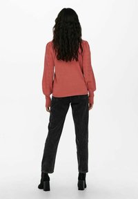 Žena s dlouhými tmavými vlasy, oblečená v rustikově zbarveném svetru s dlouhým rukávem a černými džíny s vysokým pasem, otočená zády na bílém pozadí.