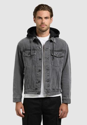 Młody mężczyzna w szarej kurtce jeansowej z czarnym kapturem, białej koszulce i czarnych spodniach, stojący na tle jednolitego jasnego tła.