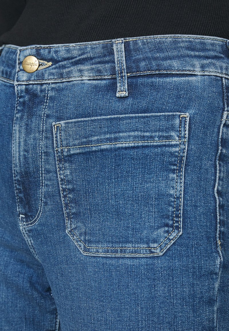 Nahaufnahme von blauen Jeans mit hinterem Taschenbereich, Gürtelschlaufe und Metallknopf am Bund, getragen mit schwarzem Oberteil.