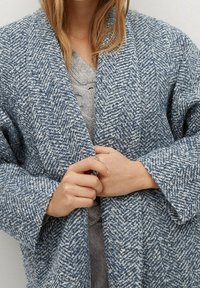Cappotto a motivi a spina di pesce blu e bianco con maniche larghe, lasciato aperto su un maglione grigio. L'attore tiene insieme i bordi frontali.