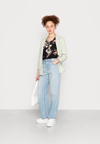 Blazer verde claro sobre uma blusa preta com padrão floral, combinado com jeans de ganga azul clara e sapatilhas brancas. Os acessórios incluem uma mala branca.