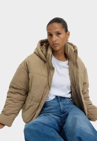 Polstret beige jakke med hætte, der har en glat tekstur og syede detaljer, båret over en hvid skjorte og blå denim jeans.