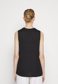 Femme aux cheveux blonds en chignon, portant un haut noir sans manches et un pantalon blanc, vue de dos sur un fond blanc uni.