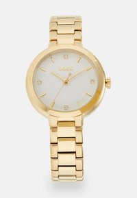 Montre-bracelet en métal doré avec un boîtier rond, un cadran blanc avec quatre marqueurs d'heure en cristal, des aiguilles dorées et un bracelet à maillons.