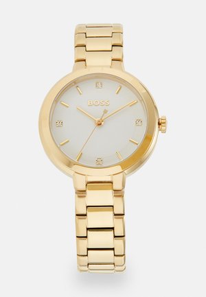 Montre-bracelet en métal doré avec un boîtier rond, un cadran blanc avec quatre marqueurs d'heure en cristal, des aiguilles dorées et un bracelet à maillons.