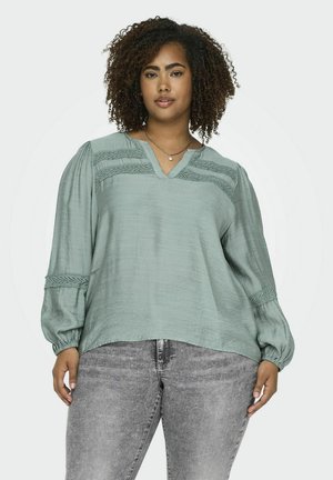 Blusa verde claro hecha de tela texturizada con escote en V, intrincados detalles de encaje en los hombros y mangas largas abullonadas.