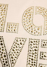 Beige textiel met gouden ronde en vierkante metalen studs die grote letters en patronen vormen, met verschillende groottes en texturen.