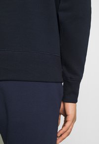 Primo piano della mano destra di una persona appoggiata al fianco, che indossa una felpa blu scuro e pantaloni abbinati su uno sfondo semplice.