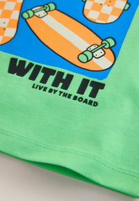 Tricou verde din bumbac, cu grafică de skateboard în portocaliu și albastru, cu text negru "WITH IT" și "LIVE BY THE BOARD".