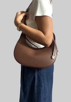 Borsa a tracolla in pelle marrone con forma curva, dotata di una sola tracolla, accenti metallici e una texture liscia. Indossata sul braccio.