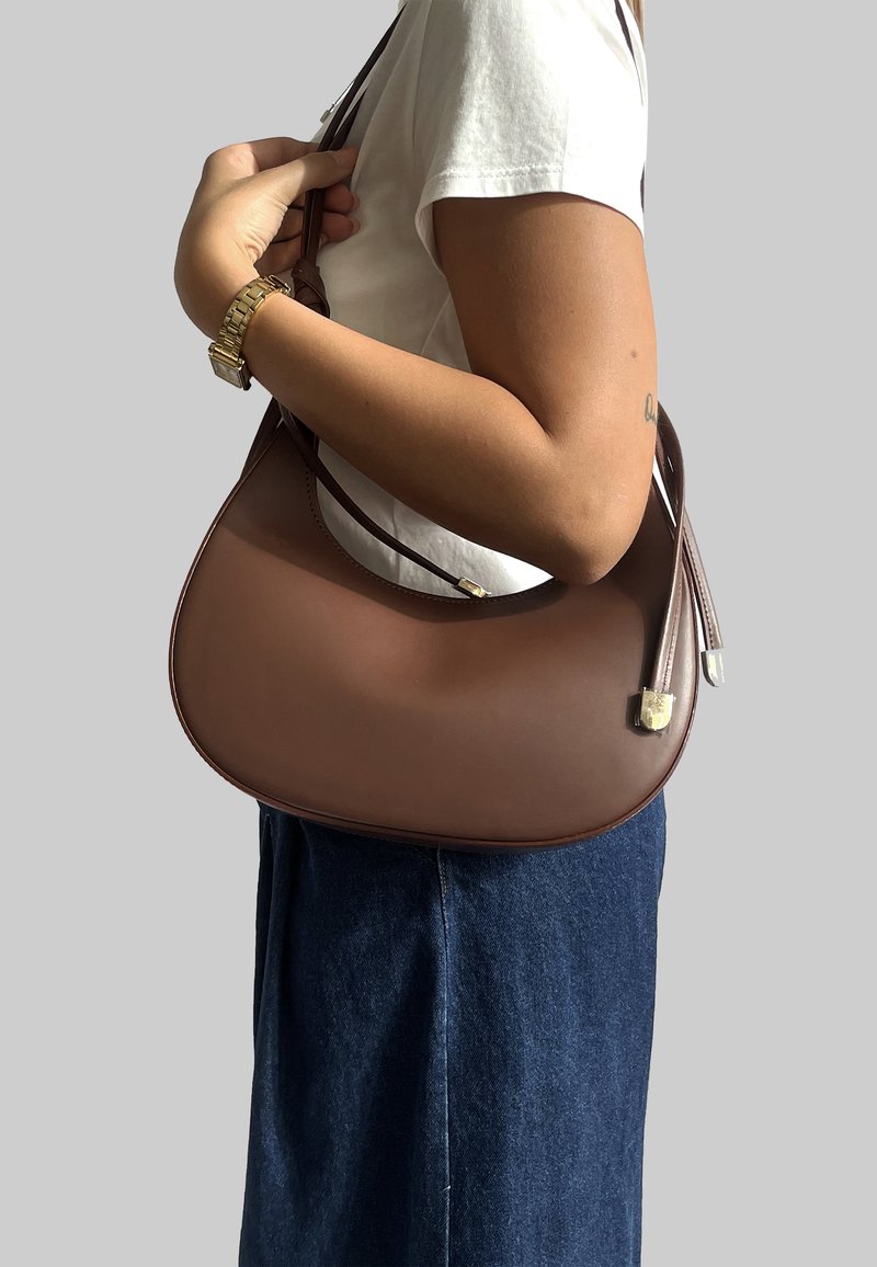 Borsa a tracolla in pelle marrone con forma curva, dotata di una sola tracolla, accenti metallici e una texture liscia. Indossata sul braccio.