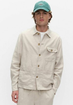 Nuori mies pukeutuneena beigen väriseen napitettuun takkiin ja housuihin, päässään vihreä lippalakki, jossa lukee "Slow Sundays Since 1974" yksinkertaisen valkoisen taustan edessä.