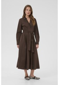 Brown belted shirt dress made of cotton, long sleeves, button-down front, and a flowing skirt. Paired with black and white striped flats.