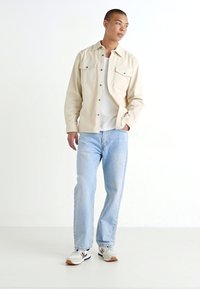 Svijetlosmeđi košulja s dugim rukavima i dva džepa na prsima, uparen s svijetloplavim trapericama i bijelim tenisicama s crvenim i mornarsko plavim detaljima.