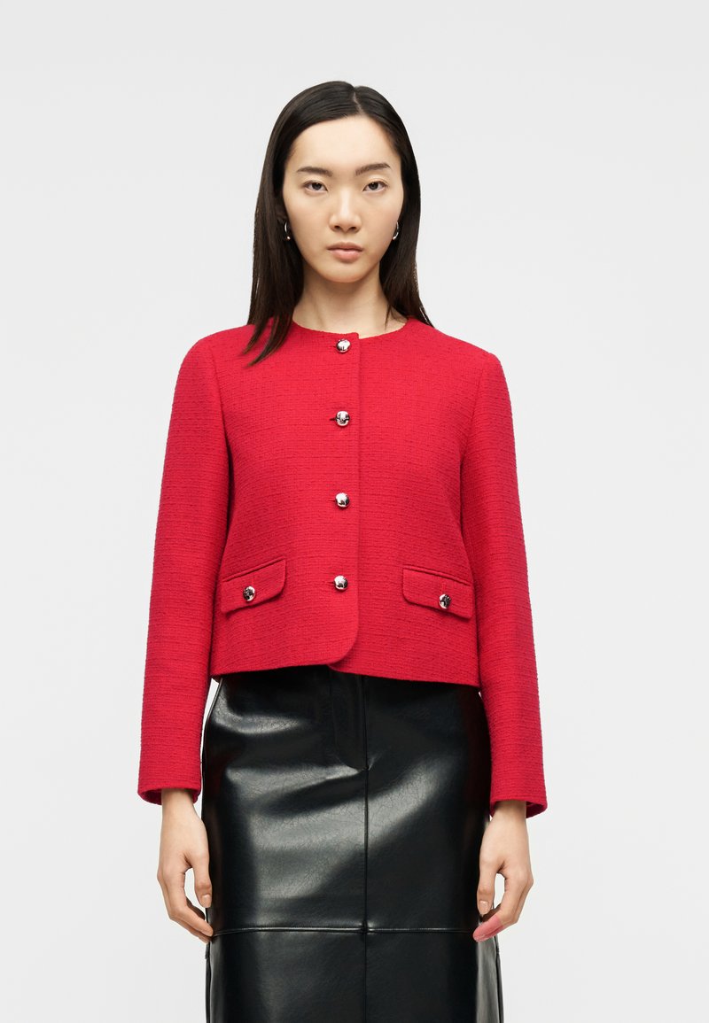 Γυναίκα με ίσια σκούρα μαλλιά που φοράει κόκκινο σακάκι με κουμπιά και τσέπες με καπάκι και μαύρη δερμάτινη φούστα, μπροστά σε απλό ανοιχτό φόντο.