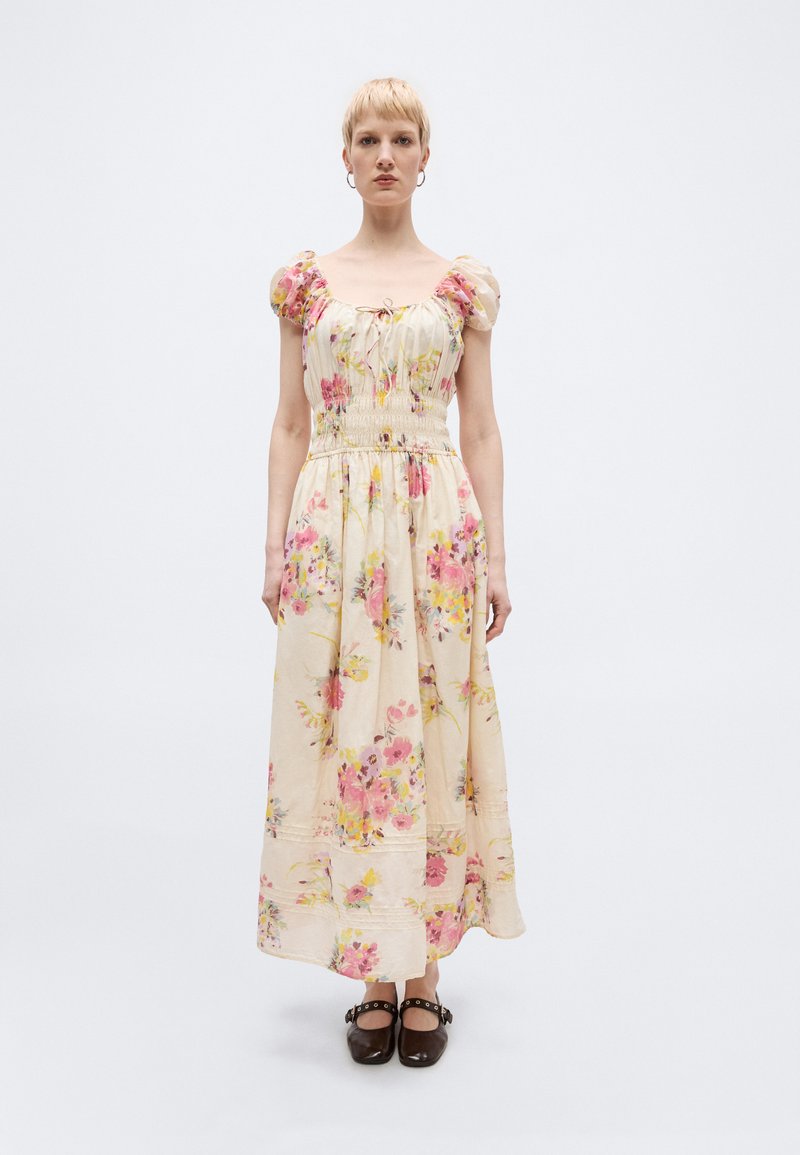 Γυναίκα με κοντά ξανθά μαλλιά, ντυμένη με μακριά μπεζ φόρεμα με φλοράλ σχέδιο και φουσκωτά μανίκια, φορώντας μαύρα παπούτσια τύπου Mary Jane, στέκεται μπροστά σε απλό φόντο.