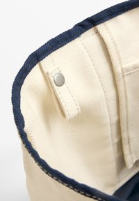 Platnena torba v kremni barvi z mornarsko modrim obrobom, opremljena s srebrnim gumbom za zapiranje, vidnimi šivi in teksturirano notranjostjo.