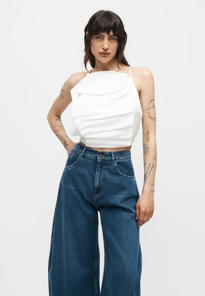 Άτομο με σκούρα μαλλιά και τατουάζ φοράει λευκό πλισέ αμάνικο τοπ και μπλε τζιν με ψηλή μέση και φαρδιά μπατζάκια σε απλό φόντο.