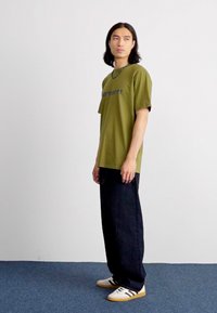 Olivgrünes Baumwoll-T-Shirt mit "Carhartt"-Logo, kombiniert mit lockeren, dunklen Jeans und weißen Sneakers mit schwarzen Streifen. Einfaches, lässiges Design.