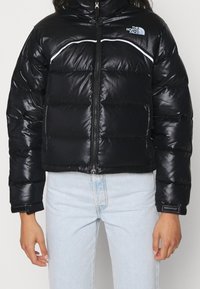 Black puffer jacket with high collar, glossy finish, white accents, and front zipper. Logo on upper left chest. Features a cropped design.