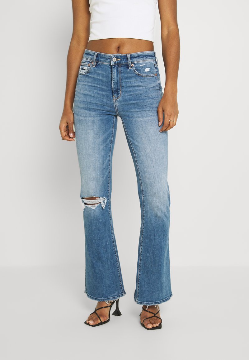 Calças de ganga azul de boca larga com uma cintura de altura média e detalhe de joelho desgastado. Apresenta um efeito desbotado, bainha desfiada e um design clássico de cinco bolsos.