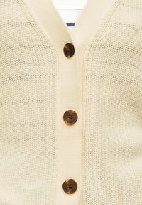Κρεμ πλεκτή ζακέτα με τρία καφέ κουμπιά, φορεμένη πάνω από λευκό και ναυτικό ριγέ τοπ, που δείχνει λεπτομέρειες υφής υφάσματος.