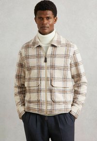 Mand iført beige og brun ternet zip-jakke over en hvid rullekrave sweater med mørke plisserede bukser, hænderne i lommen.