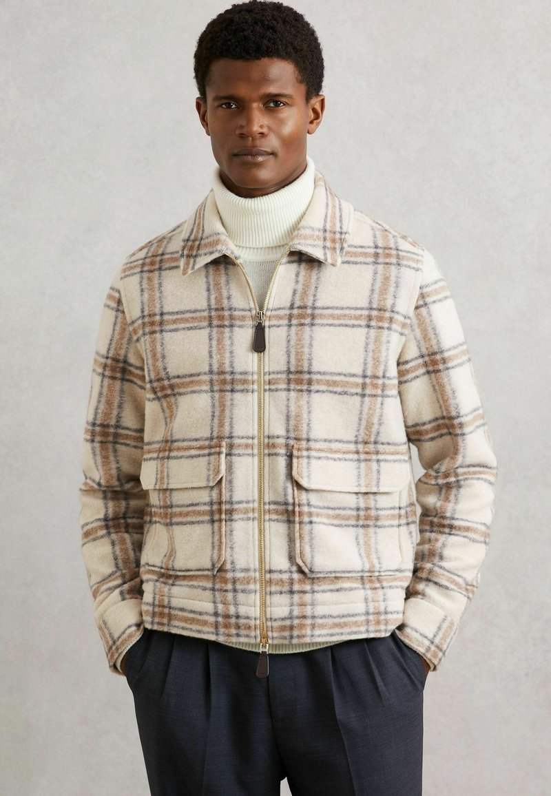 Mand iført beige og brun ternet zip-jakke over en hvid rullekrave sweater med mørke plisserede bukser, hænderne i lommen.