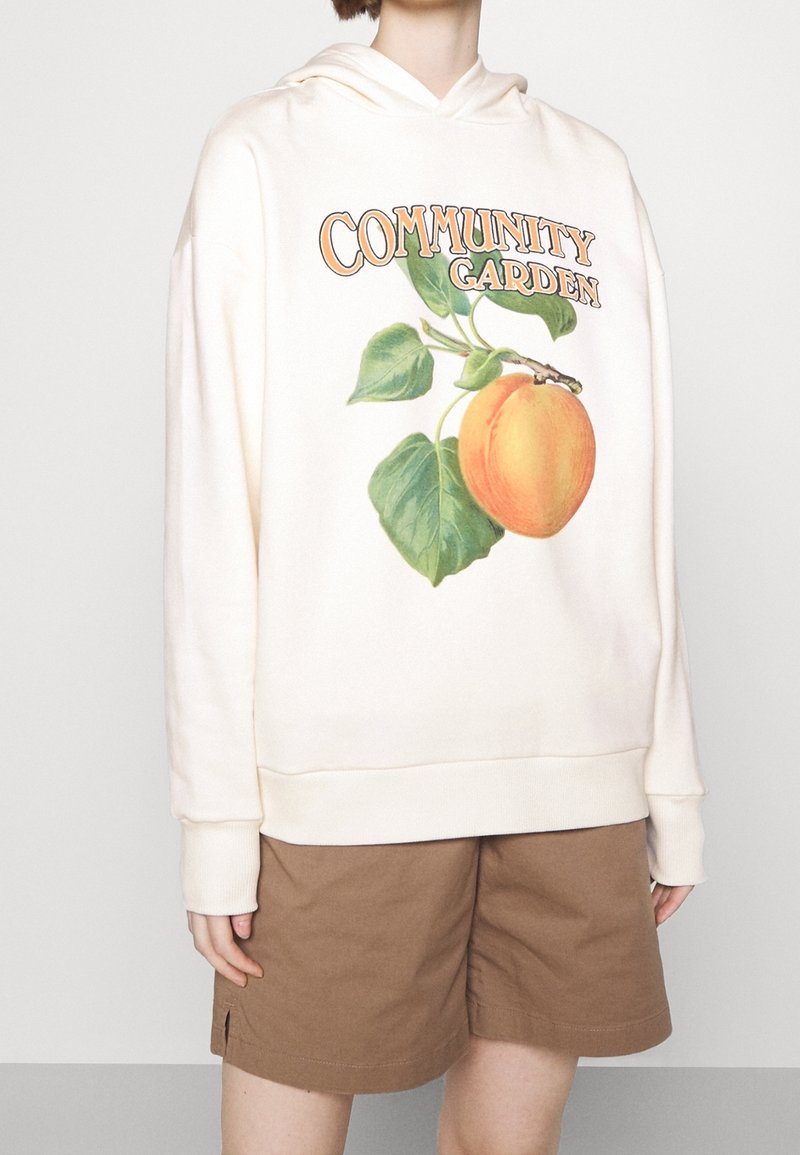 Personne portant un sweat à capuche crème avec le texte "Community Garden" et un graphisme de pêche, accompagné d'un short marron clair sur un fond uni.