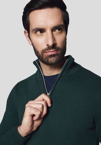 Ciemnozielony sweter z dzianiny z zamkiem błyskawicznym o ćwierć długości, z prążkowaną teksturą; posiada metalowy zamek i granatową koszulę pod spodem. Model trzyma zamek jedną ręką.