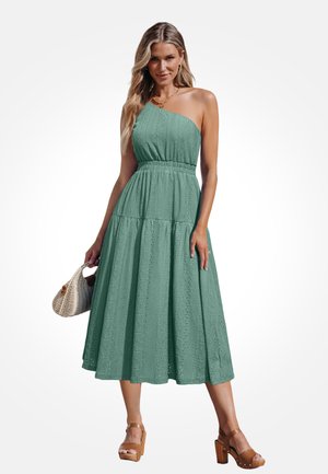 Mujer con vestido midi verde de un solo hombro, sosteniendo un bolso tejido, con sandalias de tacón alto color camel, frente a un fondo blanco.