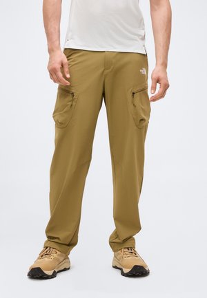 Άνδρας που φοράει μπεζ παντελόνι cargo με φερμουάρ στις τσέπες και μπεζ παπούτσια πεζοπορίας, στέκεται σε απλό φωτεινό φόντο.