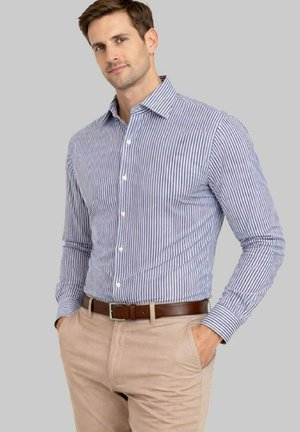 Homme portant une chemise à rayures verticales bleu et blanc à boutons, un pantalon beige et une ceinture marron, les mains dans les poches, sur un fond gris.