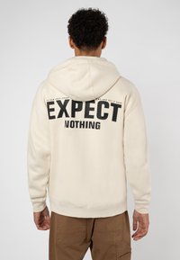 Light cream hoodie with black "EXPECT NOTHING" text on back, made from soft fabric, featuring a drawstring hood and ribbed cuffs.