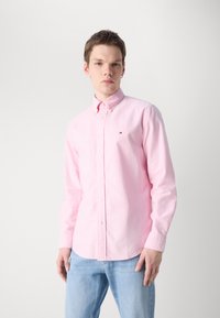 Gaiši rozā krekls ar garām piedurknēm, apkakli un mazu logo uz krūšu daļas, kopā ar gaiši ziliem džinsiem. Gluda tekstūra.