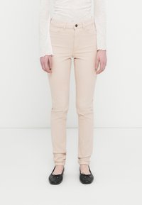 Γυναίκα που φοράει εφαρμοστά ανοιχτό μπεζ παντελόνια, λευκή μπλούζα μακρυμάνικη από δαντέλα και μαύρα μπαλαρίνες, στέκεται μπροστά σε απλό λευκό φόντο.